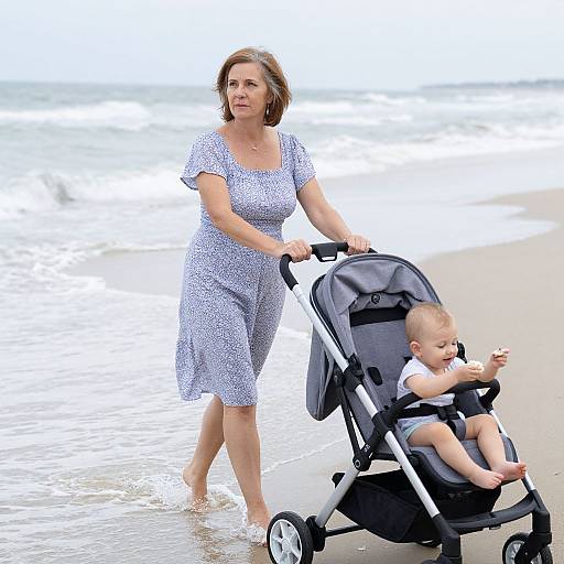 Photograph of a middle-aged woman with short brown hair in a blue floral dress, pushing a black and white baby stroller along a sandy beach with