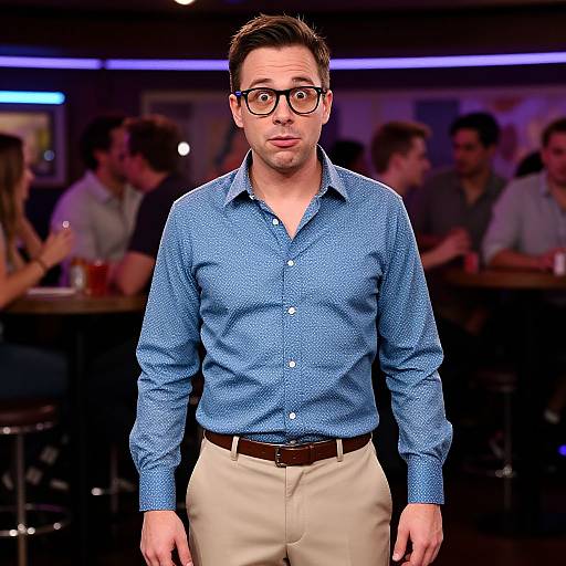 Photograph of a man with short brown hair, glasses, blue patterned shirt, beige pants, and brown belt, standing in a dimly lit