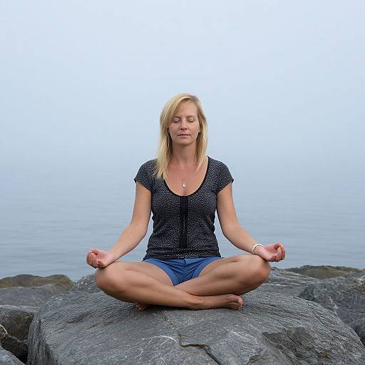 Photograph of a blonde woman in a black polka dot shirt and blue shorts, sitting cross-legged on a rocky shore in meditation pose, with calm
