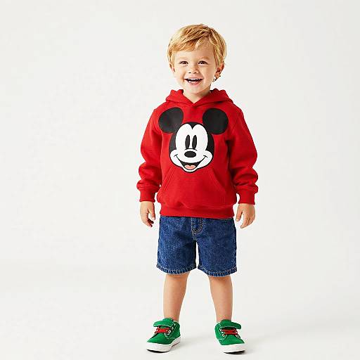 Photograph of a smiling blond toddler in a red Mickey Mouse hoodie, blue denim shorts, and green sneakers, standing against a white background.