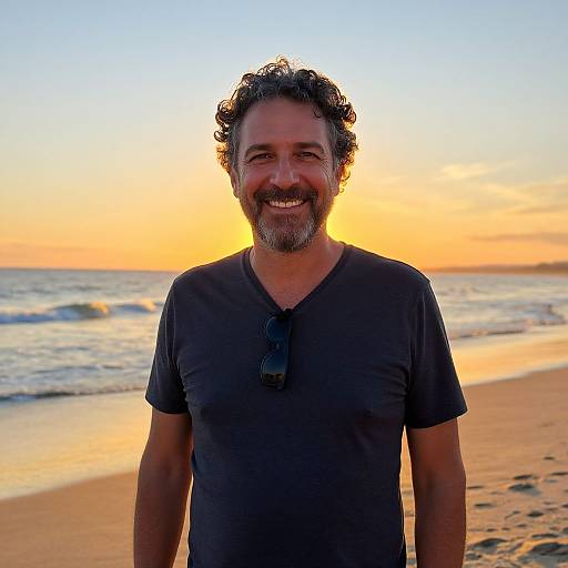 Photograph of a smiling, curly-haired, bearded man in a black t-shirt standing on a beach at sunset, with ocean waves and a vibrant