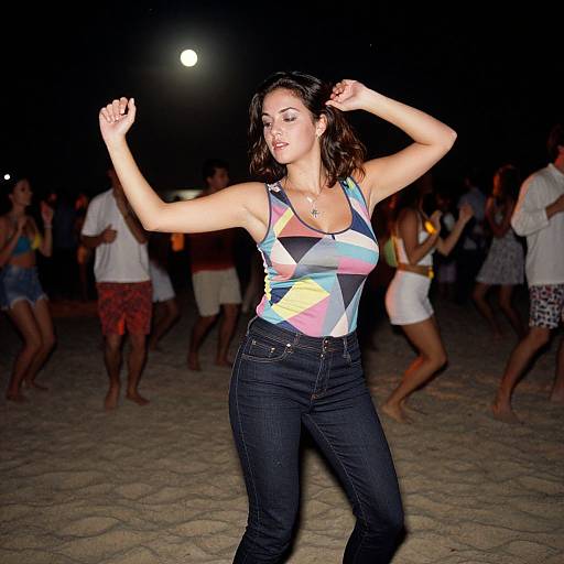 Photograph of a confident, dark-haired woman in a colorful geometric tank top and black jeans dancing at night on a sandy beach, surrounded by other dancing