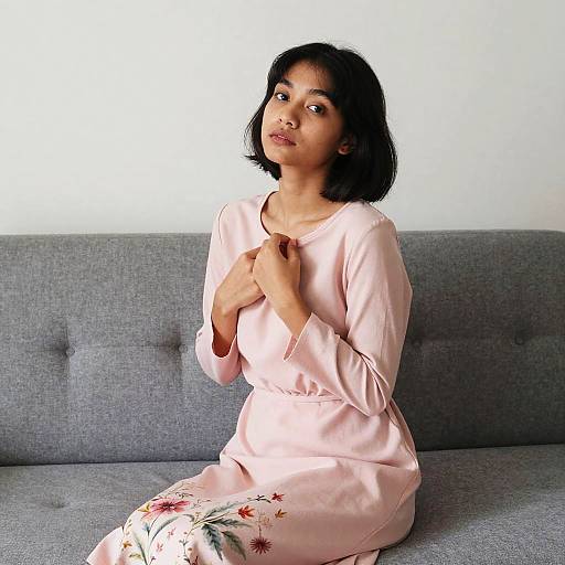Photograph of an Asian woman with short black hair, wearing a pale pink dress with floral patterns, sitting on a gray couch, hands gently touching her