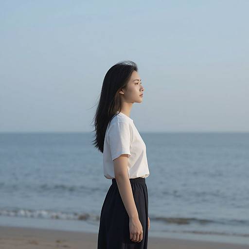 Photograph of an East Asian woman with straight black hair, wearing a white short-sleeve top and black skirt, standing in profile on a beach