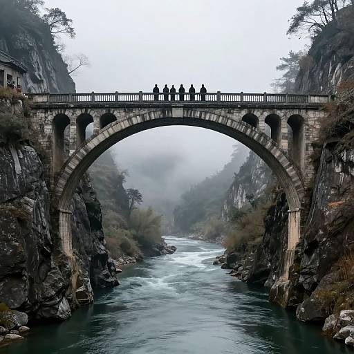 Photograph of a stone arch bridge spanning a misty, rocky river gorge, with seven silhouetted figures standing at the bridge's center.
