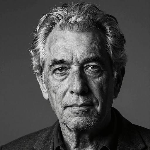 Black-and-white photograph of an elderly man with gray, wavy hair and deep-set eyes, wearing a dark jacket, against a gradient gray background.