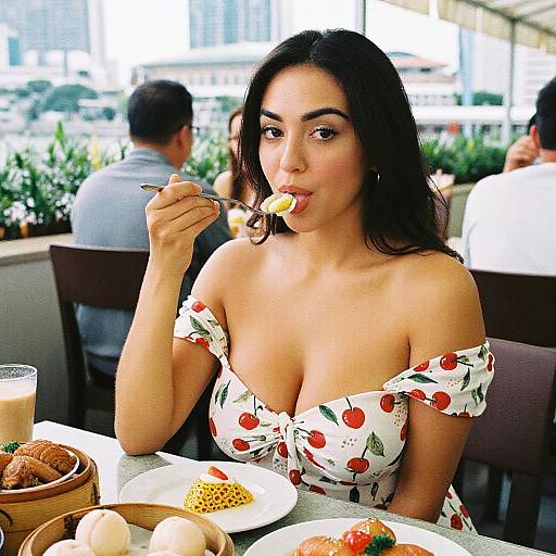 Photograph of a woman with dark hair, wearing a white off-shoulder cherry-patterned dress, eating a dessert at an outdoor café.