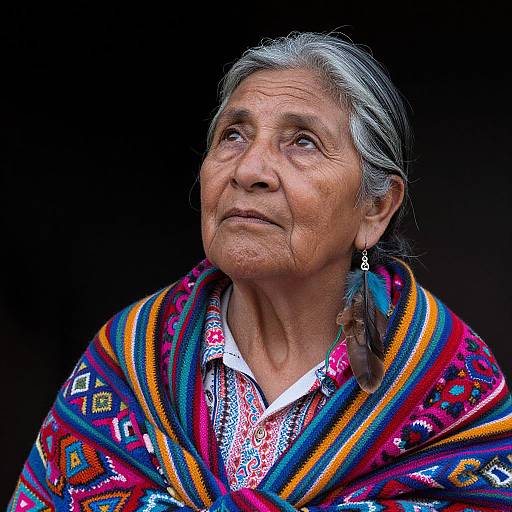 Photograph of an elderly Indigenous woman with gray hair, wearing a colorful, patterned shawl, and traditional embroidered blouse, looking upwards with a contempl
