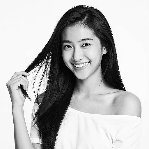 Black-and-white photograph of a smiling young Asian woman with long, straight black hair, holding a lock of it, wearing an off-shoulder white