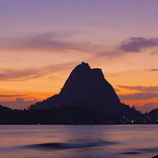 Photograph of a silhouetted, jagged mountain against a vibrant sunset sky with orange, purple, and pink hues, reflected in a calm