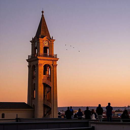 Photograph of a sunlit, Gothic-style clock tower at sunset, with silhouetted people in the foreground, against a gradient sky of orange