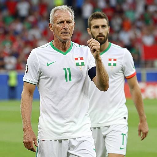Photograph of two male soccer players in white jerseys with green and red accents, standing on a grass field, one raising his fist in celebration. Bl