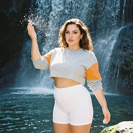 Photograph of a curvy woman with wavy brown hair, wearing a gray crop top with orange sleeves and white shorts, standing in front of a