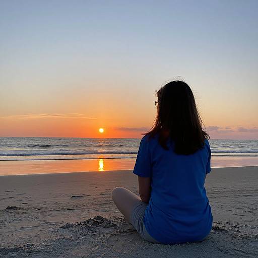 Photograph of a woman with long brown hair, wearing a blue shirt and white pants, sitting on a sandy beach, watching an orange sunset over the