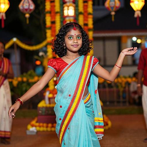 Photograph of a young South Asian girl with curly black hair, wearing a blue saree with red and yellow borders, performing a traditional dance in a