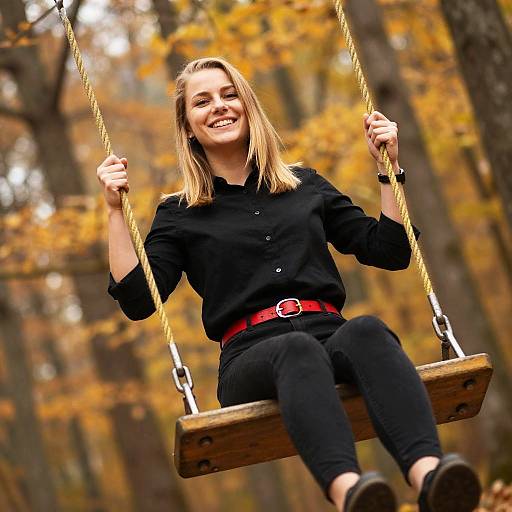 Photograph of a smiling blonde woman with straight hair, wearing a black shirt and pants, red belt, and black shoes, swinging on a wooden swing