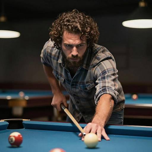 Photograph of a bearded man with curly hair, wearing a plaid shirt, intensely aiming a pool cue to strike a ball on a blue pool