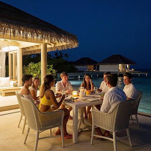Photograph of seven adults, five men and two women, dining under a thatched roof at night by a glowing pool. Wearing white outfits,