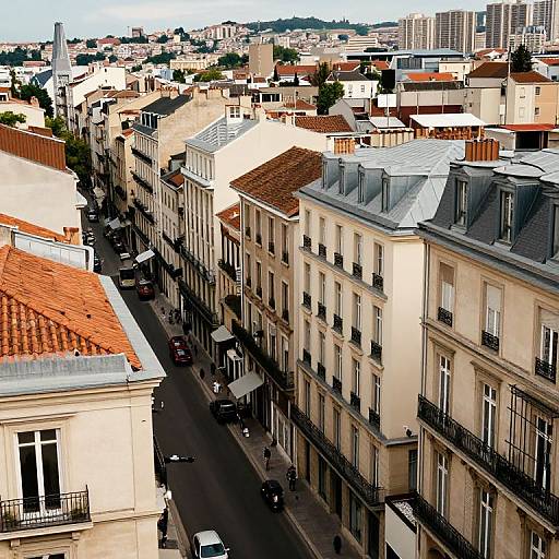 Aerial photograph of a European urban street with beige buildings, red-tiled roofs, black wrought iron balconies, and parked cars on the narrow street