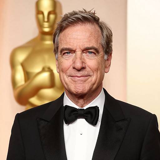 Photograph of a smiling middle-aged man with short gray hair, wearing a black tuxedo and bow tie, standing in front of a golden Oscar