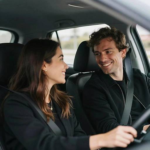 Photograph of a smiling man with curly brown hair in a black shirt, seated in the passenger seat, and a woman with dark hair in a black