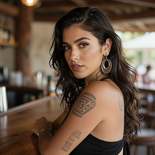 Photograph of a beautiful woman with long dark hair, hoop earrings, and tattoos, sitting at a wooden bar, looking confidently at the camera. Warm