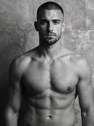 Black-and-white photograph of a muscular, shirtless, bearded man with short hair, staring intensely at the camera against a textured gray background.
