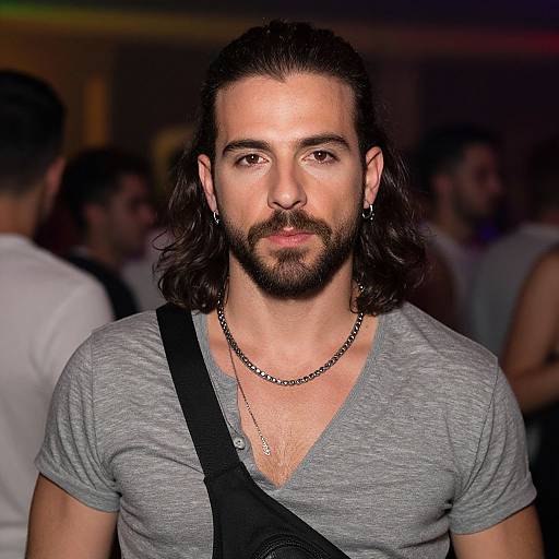 Photograph of a bearded man with long dark hair, wearing a gray V-neck shirt, black necklace, and shoulder strap, in a dimly