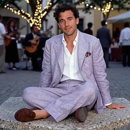 Photograph of a handsome, dark-haired man in a light purple, textured suit, white shirt, and brown shoes, sitting on a stone ledge in