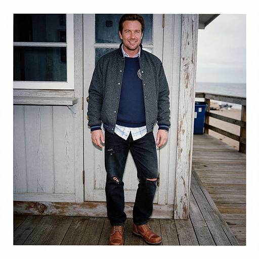 Photograph of a smiling man with short brown hair, wearing a dark grey jacket, black ripped jeans, brown boots, and a navy sweater, standing