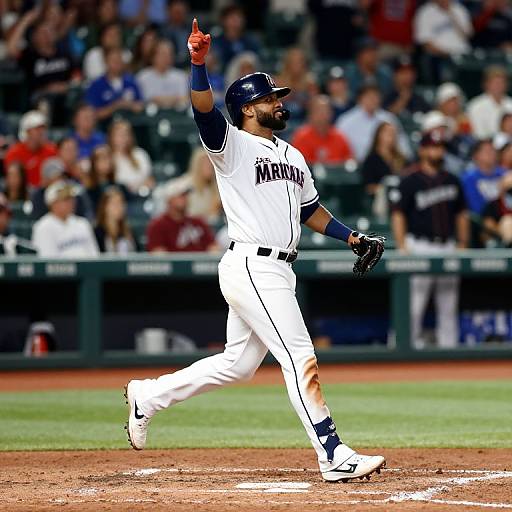 Photograph of a bearded baseball player in white Mariners uniform, navy helmet, and blue wristband, raising right index finger while running on field,