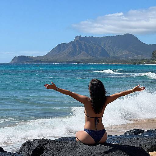 Photograph of a woman in a black bikini, arms outstretched, sitting on black rocks, overlooking ocean waves and mountains under a clear blue sky