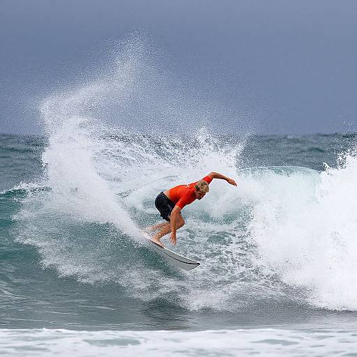 Photograph of a male surfer in a red shirt and black board shorts, riding a breaking ocean wave, with spray splashing around him.