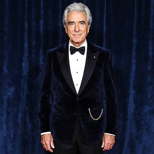 Photograph of an elderly man with gray hair, wearing a black velvet tuxedo, white shirt, black bow tie, and gold chain, standing