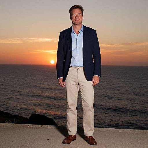 Photograph of a smiling, tall, white man with short brown hair, wearing a dark blazer, light blue shirt, beige pants, and brown