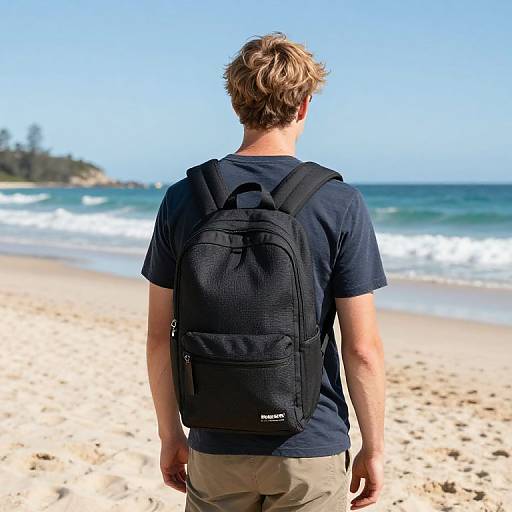 Photograph of a man with blond hair, wearing a black backpack and navy t-shirt, standing on a sunny beach, facing the ocean.