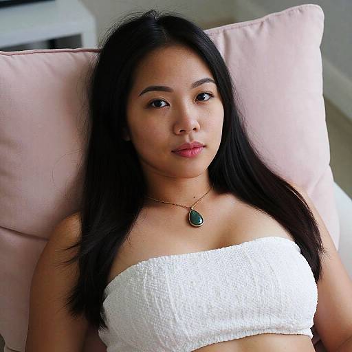 Photograph of an Asian woman with long black hair, wearing a white textured tube top and green pendant necklace, lying on a pink pillow.