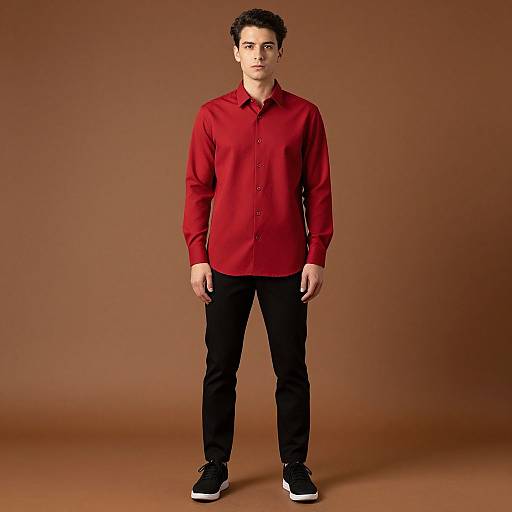 Photograph of a young man with short dark hair, wearing a red button-up shirt, black pants, and black sneakers, standing against a solid brown