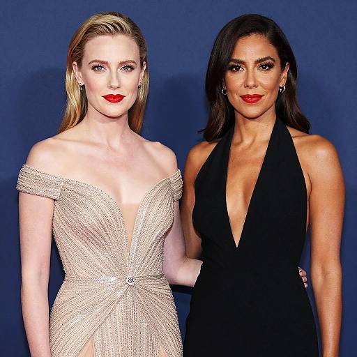 Photograph of two women in formal attire: blonde in shimmering off-shoulder gown, dark-haired in black halter dress, both with red