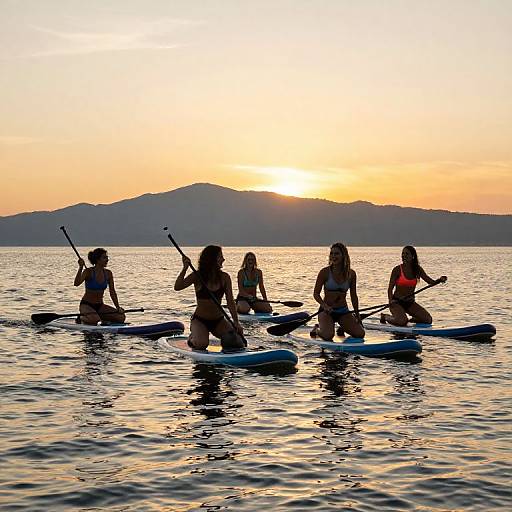 Photograph of five silhouetted women in bikinis paddleboarding on calm water at sunset, with a mountain range in the background.