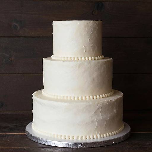 Photograph of a three-tiered white wedding cake with textured frosting and pearl-like bead decorations on each tier, set against a dark wooden background.