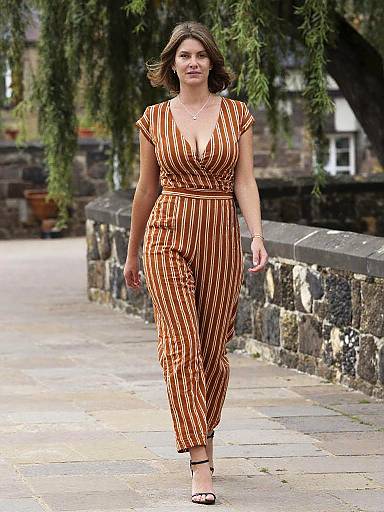 Photograph of a confident woman with wavy brown hair, wearing an orange and white striped wrap dress and white sandals, walking on a stone path with