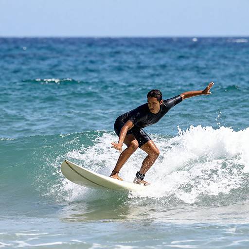 Photograph of a male surfer in a black wetsuit riding a small wave in bright blue ocean water, with clear skies in the background.