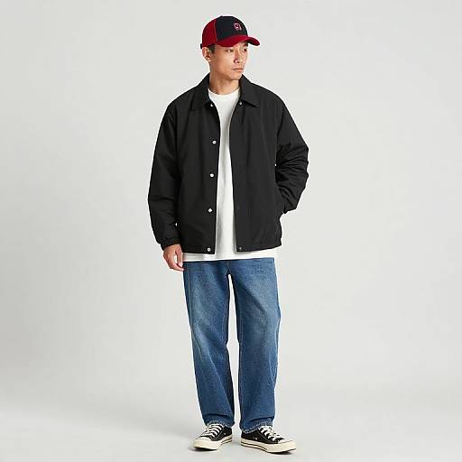 Photograph of a young Asian man in a black jacket, white shirt, blue jeans, black and white sneakers, and a red cap, standing against