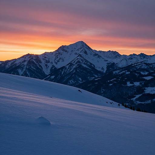 Photograph of a snow-covered mountain range at sunset, with vibrant orange and purple skies, dark forested slopes, and a smooth, blue-ting