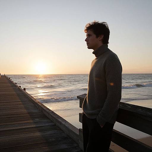 Photograph of a young man with curly hair, wearing a gray turtleneck sweater, standing on a wooden pier at sunset, gazing at the