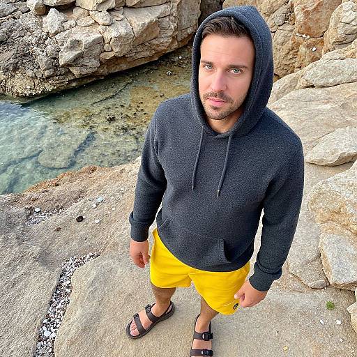 Photograph of a bearded man with brown hair, wearing a black hoodie, yellow shorts, and sandals, standing by a rocky stream.
