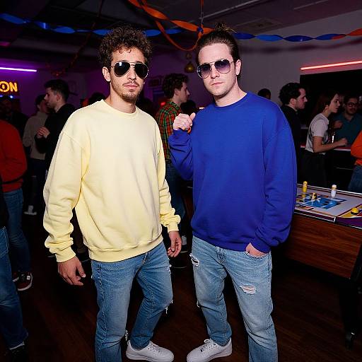 Photograph of two stylish men at a party, wearing sunglasses, yellow and blue sweatshirts, ripped jeans, and white sneakers, standing confidently.