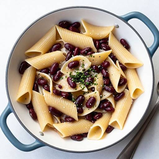 Photograph of creamy penne pasta with dark kidney beans, topped with chopped parsley, in a white enamel pot with blue handles.