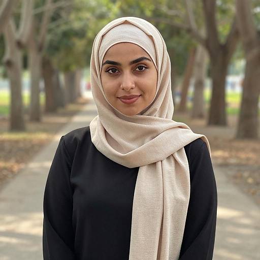 Photograph of a young woman with medium-brown skin, brown eyes, wearing a white hijab and black long-sleeve top, smiling gently